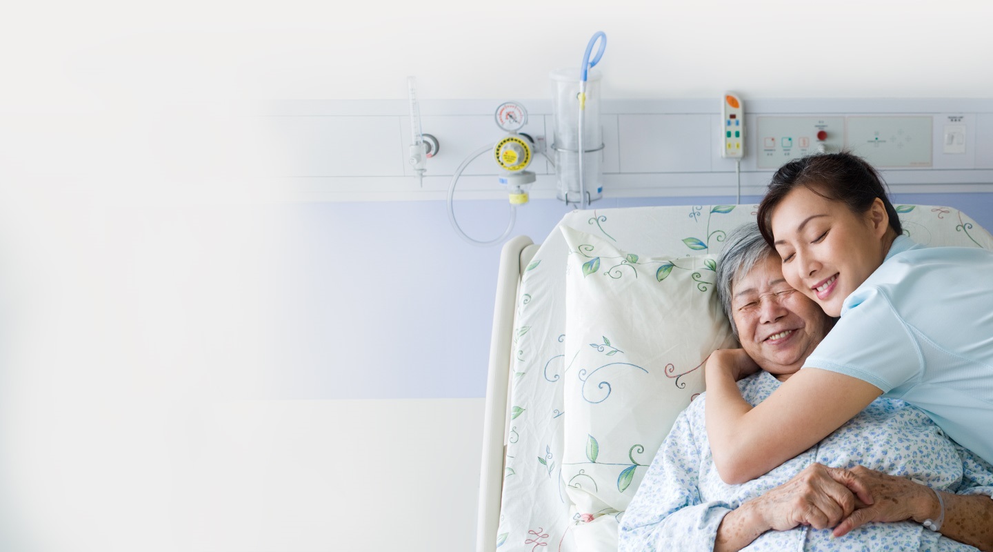 Νοσηλεύτρια που αγκαλιάζει μια ασθενή ξαπλωμένη στο κρεβάτι της στο νοσοκομείο – και οι δύο χαμογελούν.