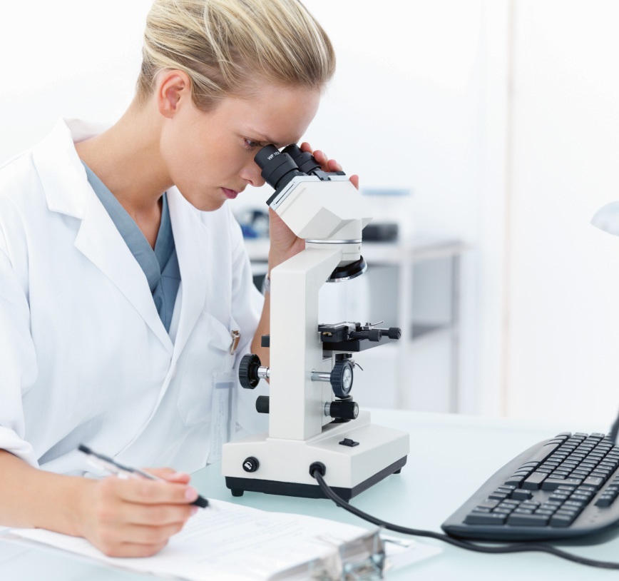 A woman, dressed in a surgery gown, is sitting in a lab, looking into a microscope