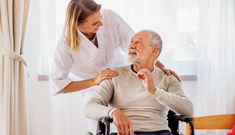 Pflegerin beugt sich von hinten über die Schulter eines im Rollstuhl sitzenden Seniore