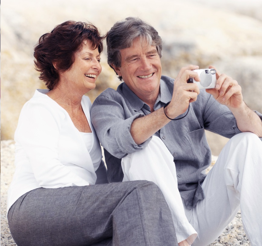 An elderly couple is sitting on rocky ground, while both of them are smiling into a camera, which the husband holds in front of them